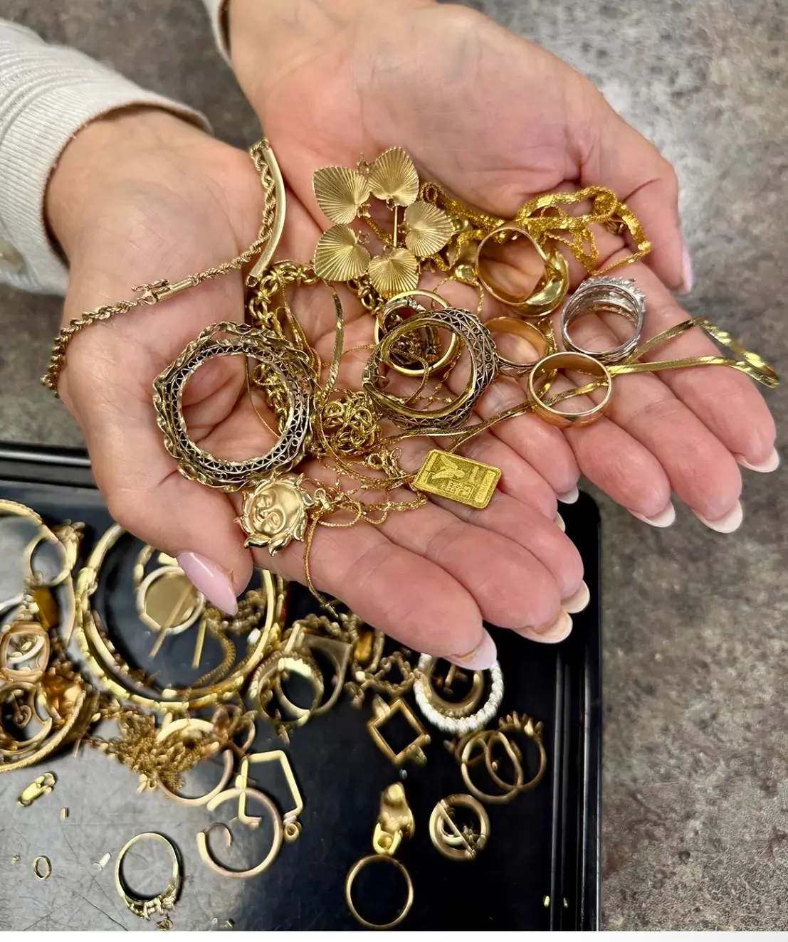 Two hands holding a pile of gold chains, rings, charms and a small gold bar over a tray of more gold jewelry.